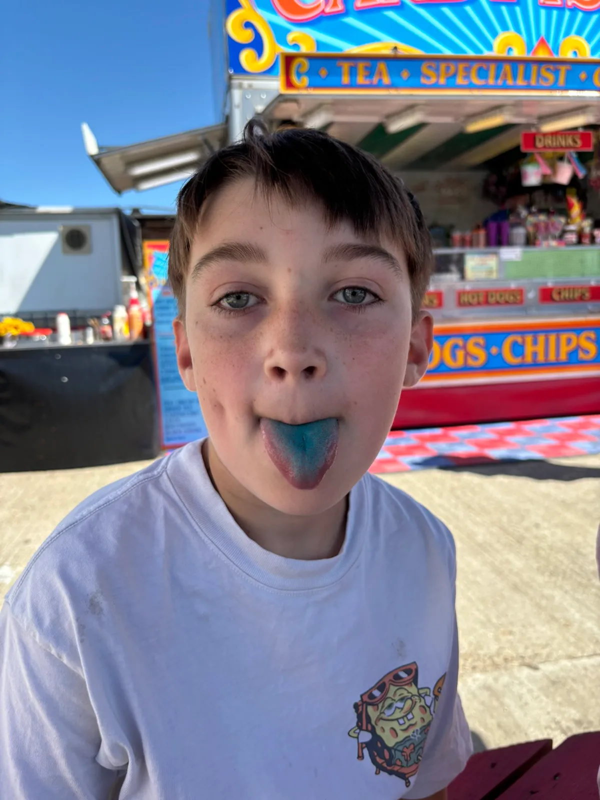 Visitor sticking out his blue tongue, coloured from the blueberry ice cream with food stall in the background.