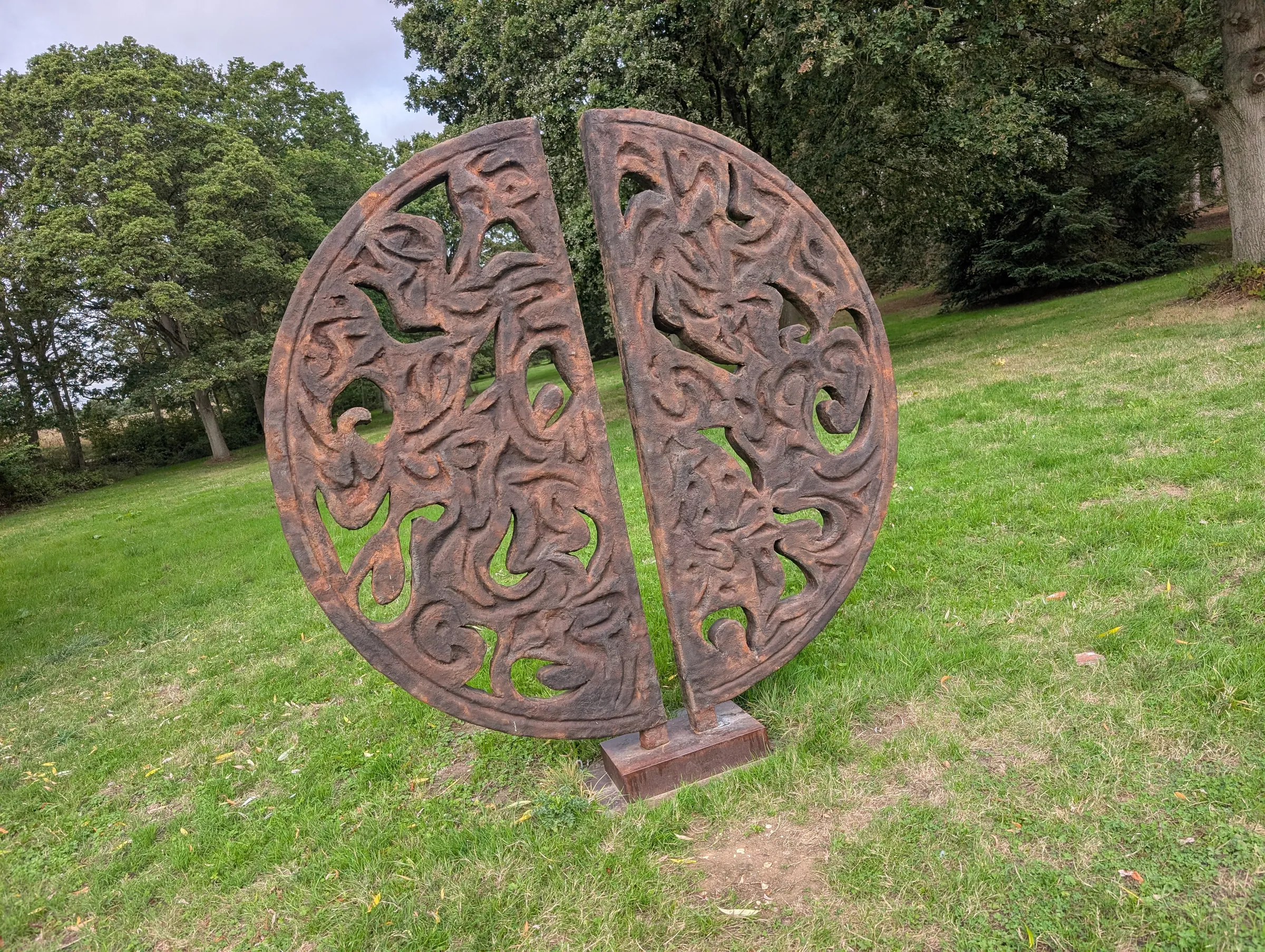 Circular metal sculpture called Benevolence among the trees and grass at Markshall Estate.